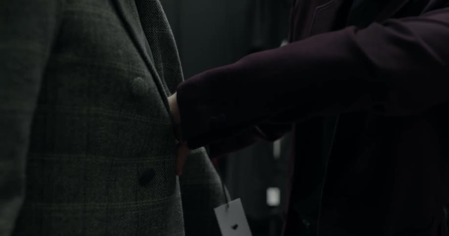 A sales person helps a man put on a suit jacket and then buttons it for him in a men's retail store