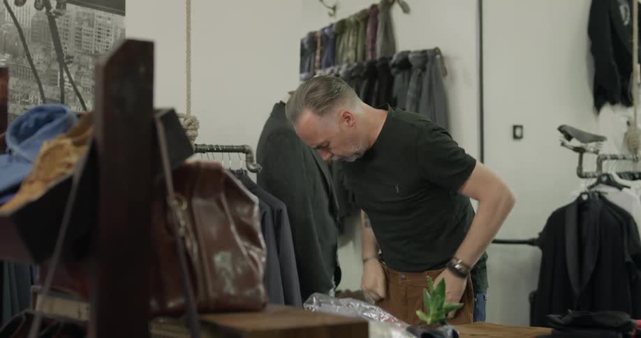 A man in a clothing store picks up a pair of brown pants and holds them against his waist