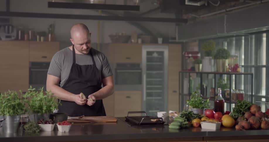 A bald man with a mustache is wearing an apron as he prepares some food and places it on a small grill that is on the same kitchen countertop that is covered in various vegetables and herbs