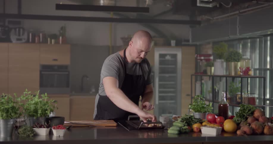 A large bald man in a dark apron is carefully turning over two pieces of meat that he is grilling on a small countertop grill within a well organized kitchen work space