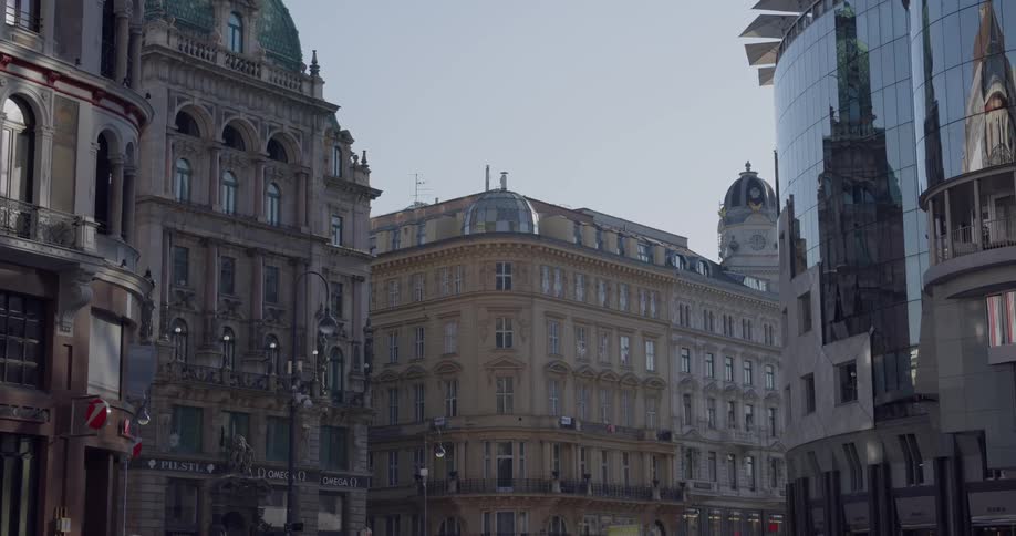 The older ornate surrounding buildings are reflected on the glass facade of a modern city structure
