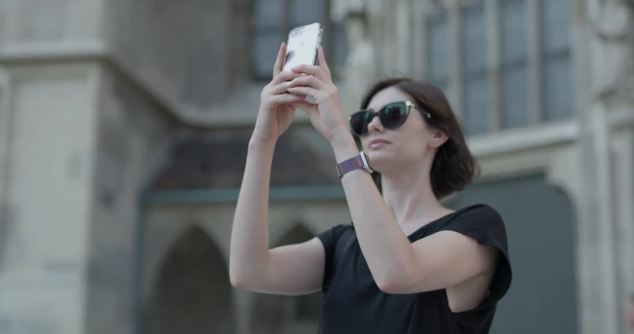 A woman walks away from a building then stops to take a picture with her phone