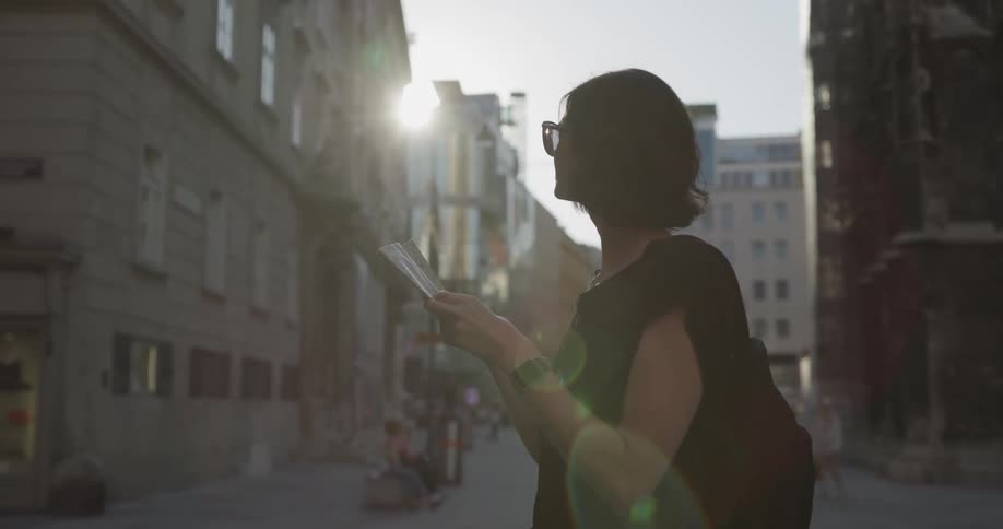 A woman in black wearing sunglasses checks a magazine while standing in a city