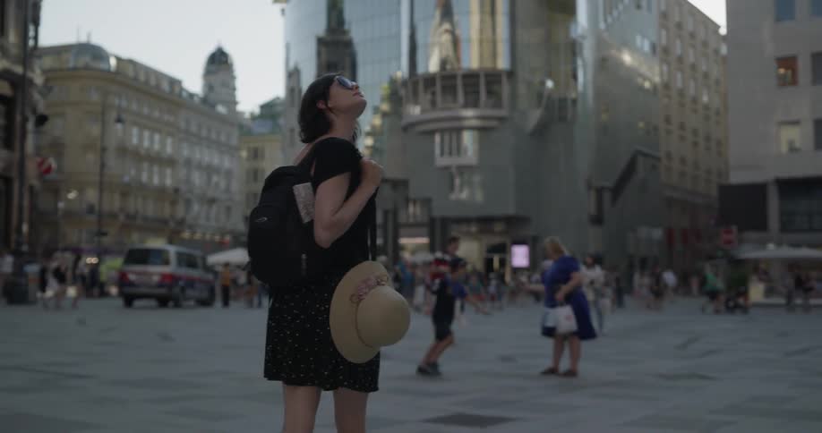 A woman carrying a sunhat and bag walks through a city plaza looking up at the buildings