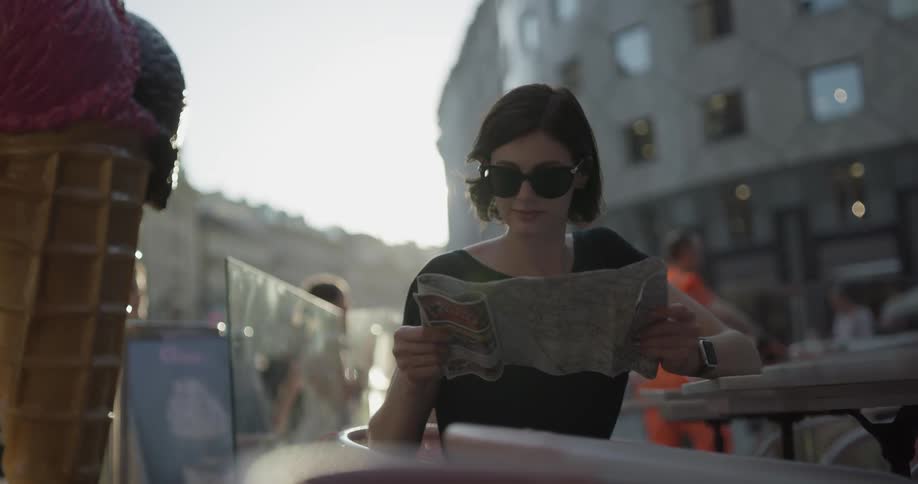 Woman wearing sunglasses sits at table and checks out a map at a cafeteria on a sunny day