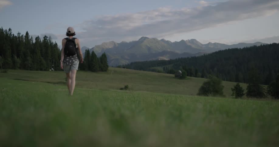 Woman wearing cap and backpack walks on the grassland with the view of distant mountains during daylight