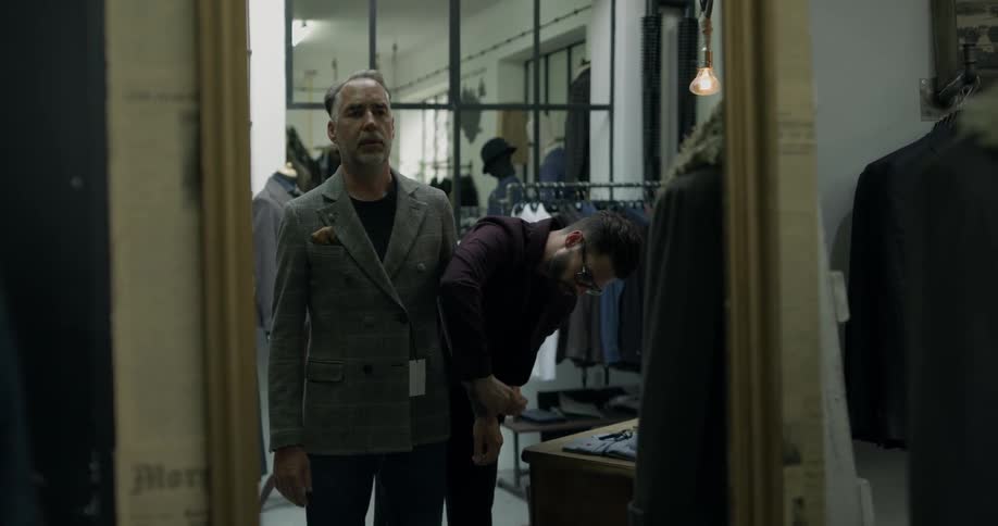 A man looking at a suit in the mirror in a clothing store while another man helps him