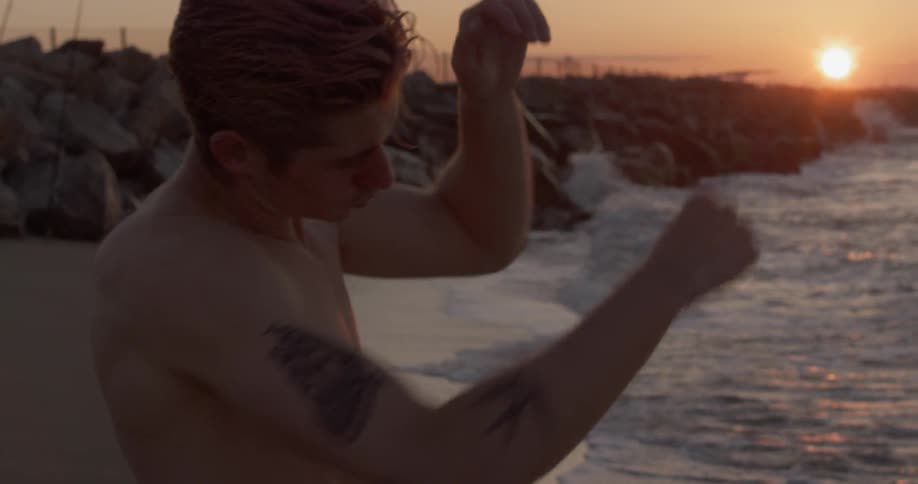 A shirtless man shadow boxing on the beach at sunset