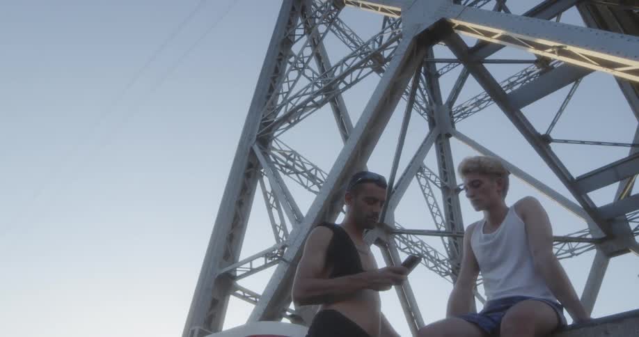 Two young men are up on electrical signal tower one is sitting and looks as if he has been crying while the other us tapping on his smartphone