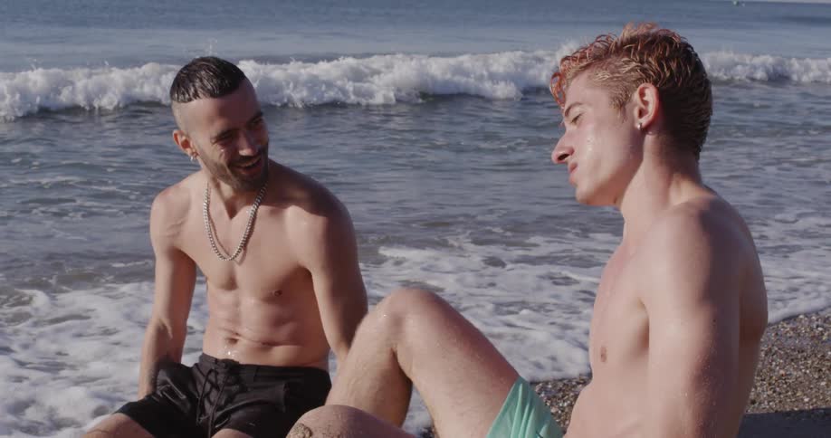 Two young white men in swim trunks sit on the beach