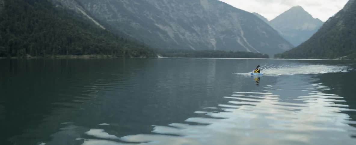 A kayaker rows across a lake surrounded by forest and mountains