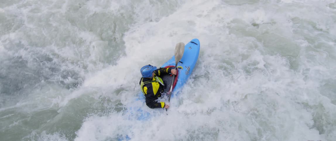 A person in a kayak moving down fast waves toward a large rock
