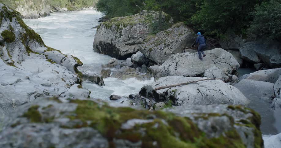 A lone person is rock hopping trying to avoid falling into the cold raging river