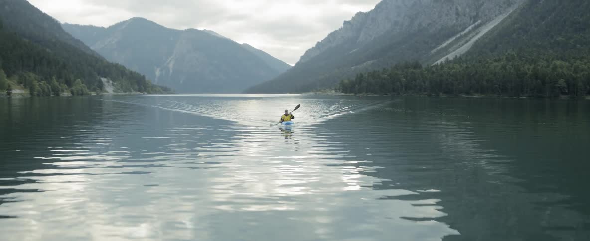 A kayaker paddles across a lake surrounded by mountains and forest