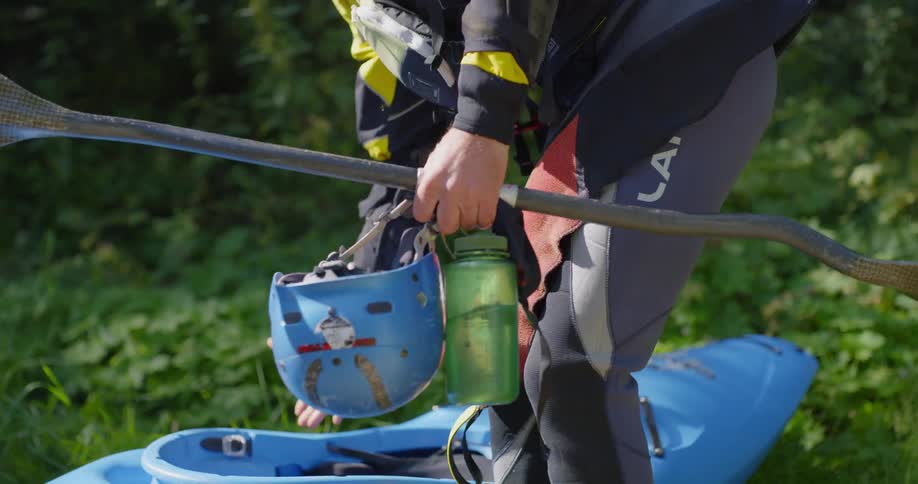 A male caucAsian in grass carrying a kayak paddle, helmet, and a water bottle