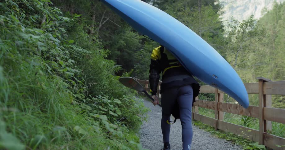 A man carries a blue kayak on his back while hiking in mountains