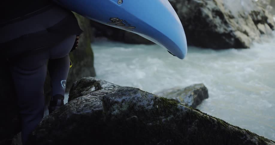 A person walks on rocks next to a river carrying a kayak
