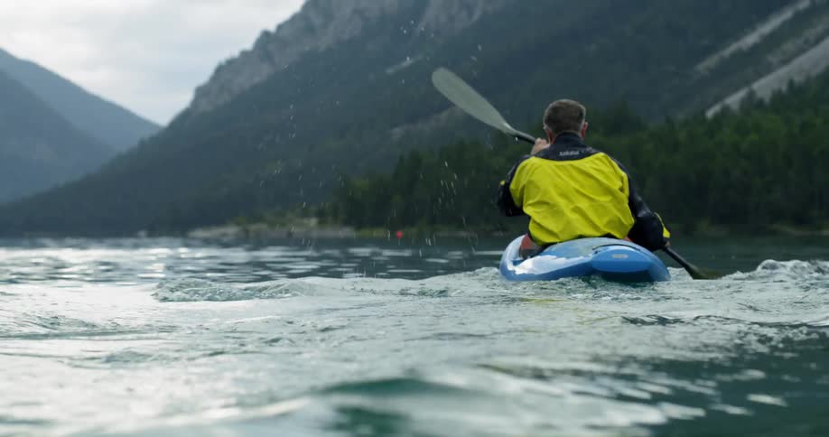A man paddles in a blue kayak on a mountain lake