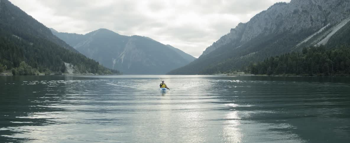 A man paddles a kayak down a river that sits between mountains