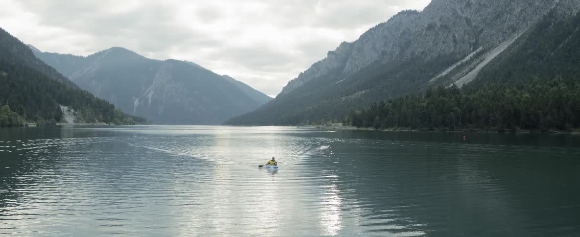 A kayaker paddles across a lake surrounded by forest and mountains