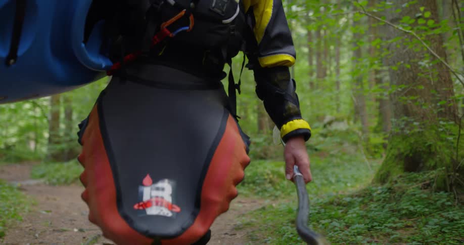 A white man walks along a forest path while carrying a kayak and an oar