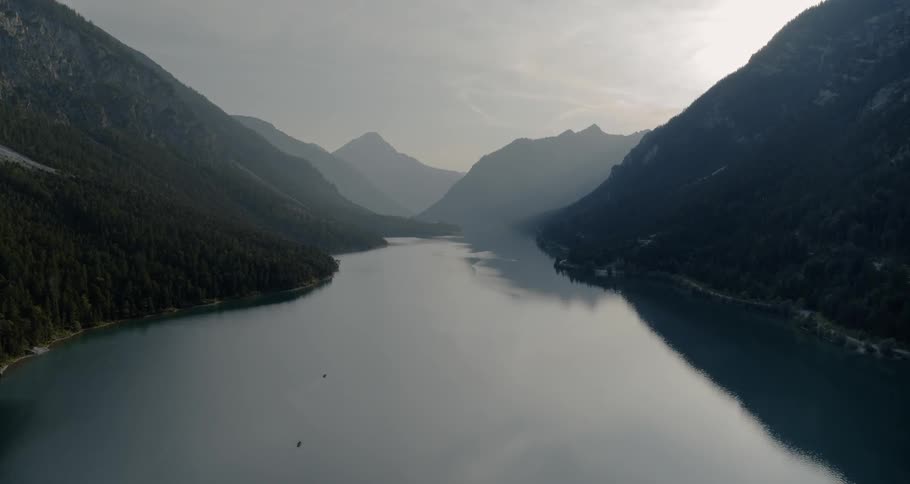A lake is surrounded by forest covered mountains while a drone is flying in the middle