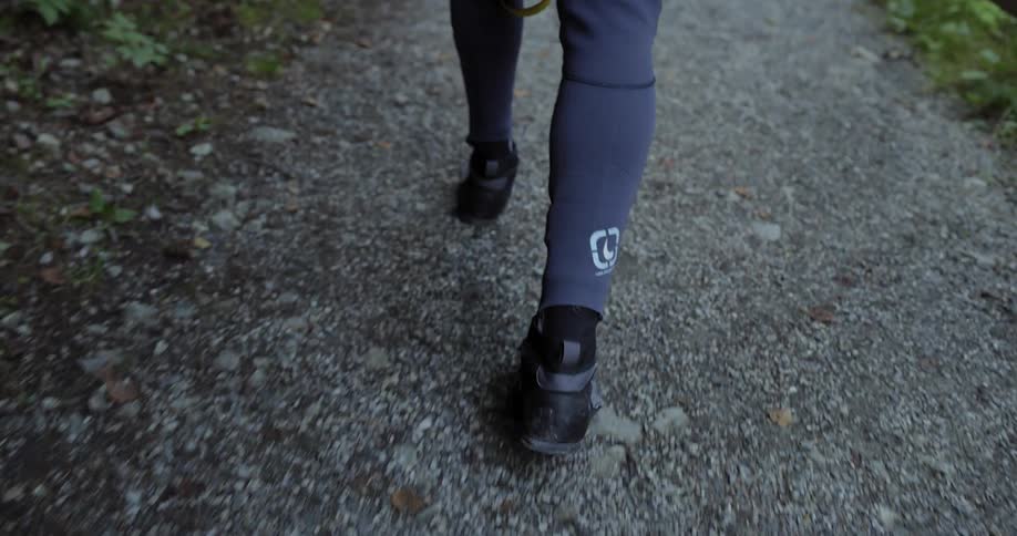 Feet wearing odd looking specialty footwear and purple clad ankles are walking down a gravel path