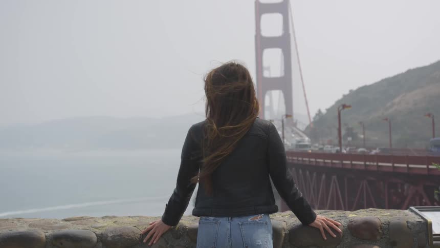 A woman stands along a stone wall next to the golden gate bridge
