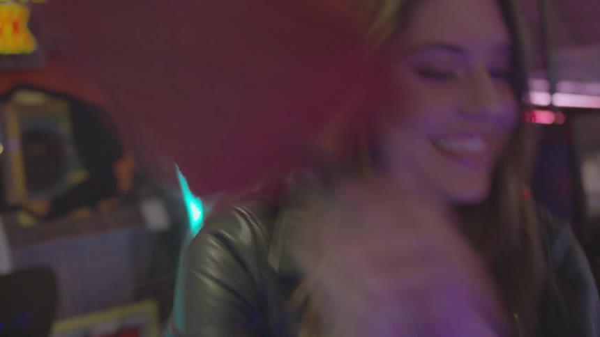 A brunette female in a leather jacket wields an oversized hammer in an arcade