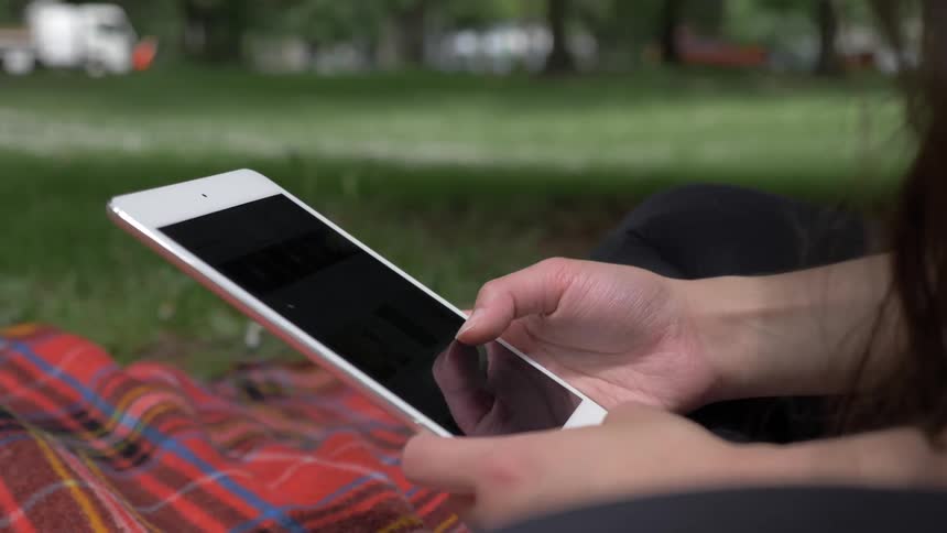 A woman scrolls on a tablet while sitting on a plaid blanket in a park