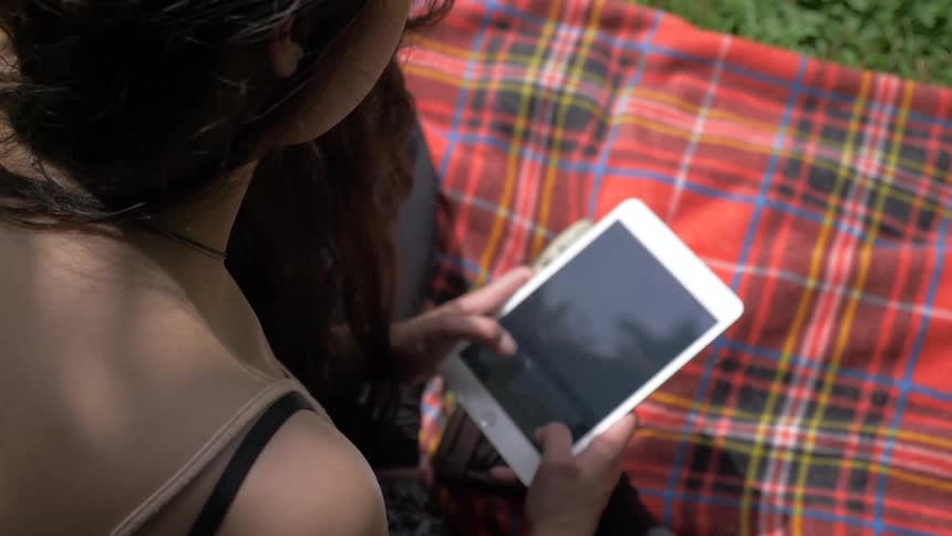 A woman typing on a tablet while sitting on a blanket in the grass