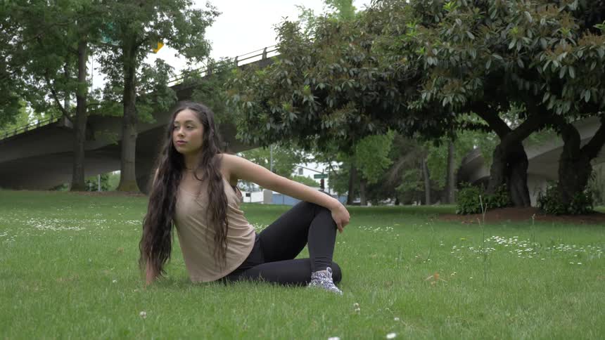 A young long haired caucasian female is sitting down stretching her body on grass