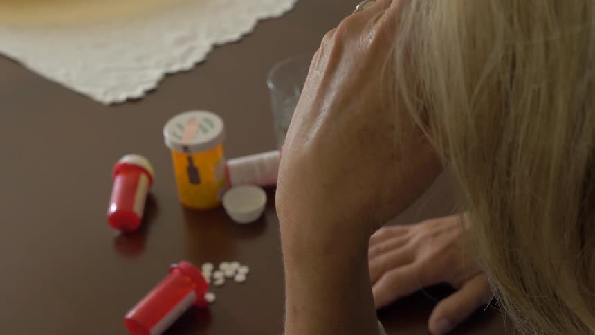 A woman with blonde hair sits at a table with her hand on her forehead as there are a few pill bottles and one bottle that is spilled with the pills scattered