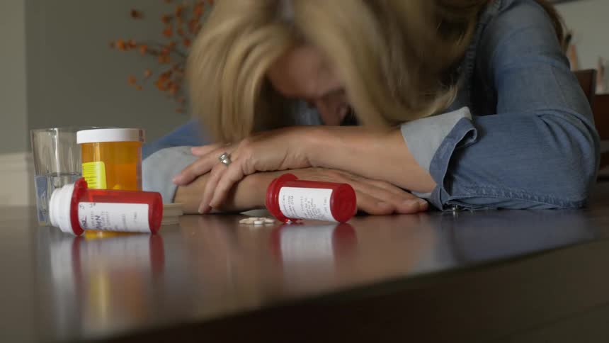 A woman puts her head on the table next to several bottles of pills