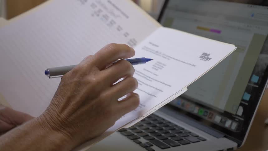 A caucasian person holding and writing in a folder near a laptop