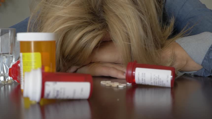 Woman signs and lowers her head on a table with pills and pill bottles on it