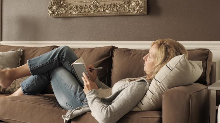 A woman lays down on a couch and writes in a journal inside