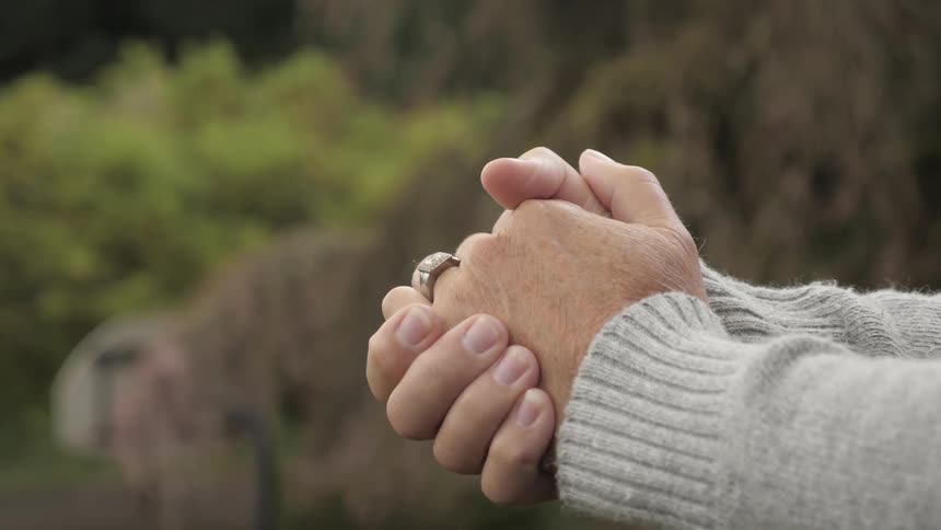 A woman wearing a beige sweater and wedding ring wrings her hands in a park setting