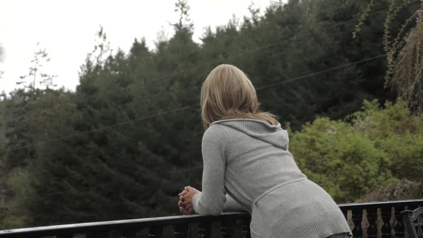 Blonde woman puts her elbows on the banister and looks at the trees on a cloudy day
