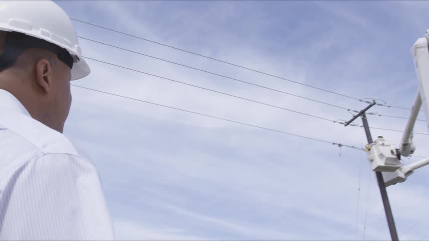 A man standing on the ground watches a worker in a cherry picker fixing a power line