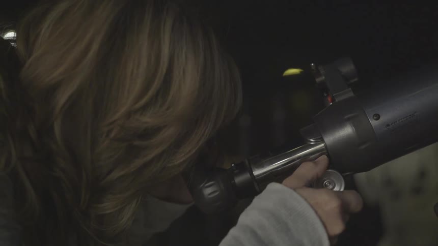 A woman bends down and looks through a telescope turning a knob to adjust the angle