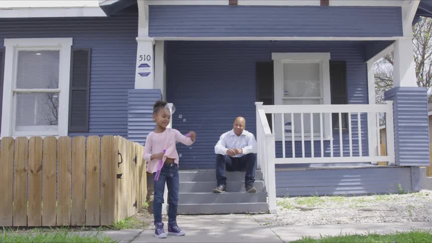 Young girl blows bubbles in front of her house while her dad sits on the steps and watches