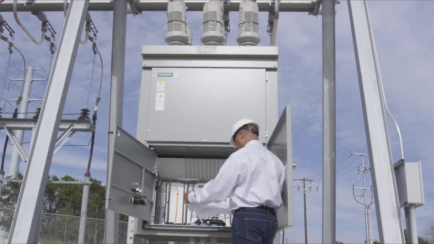 A black man in a construction hat looking at a paper while standing before an open power box