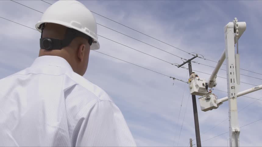 A man in a white hard hat stands on the ground looking at a man in a bucket lift working on power lines