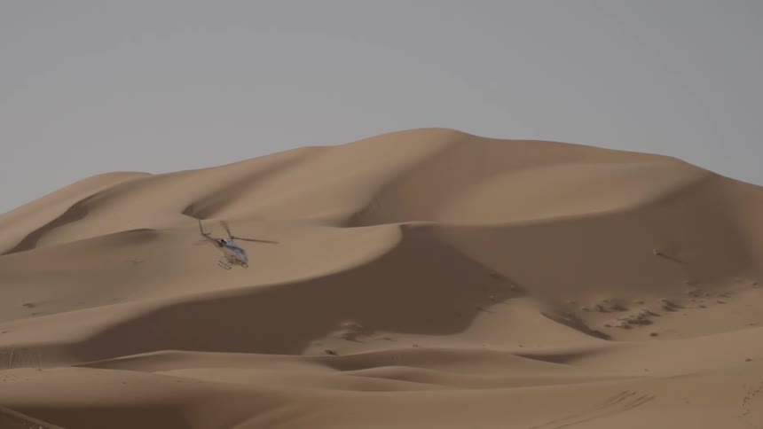 A helicopter flies by sand dunes in the desert