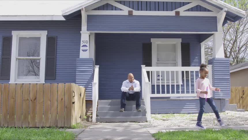 Father sits on the porch steps smiling and clapping at his daughter as she runs along the sidewalk waving a wand making bubbles appear in the air