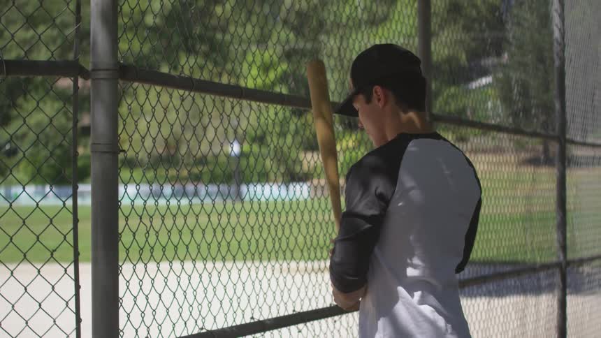 Baseball player holds the wire fence and practices with baseball bat in the daytime