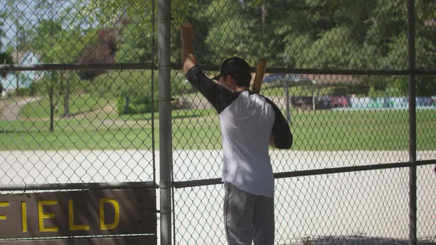 A young man wearing a ball cap and holding a wooden baseball bat looks at a baseball field