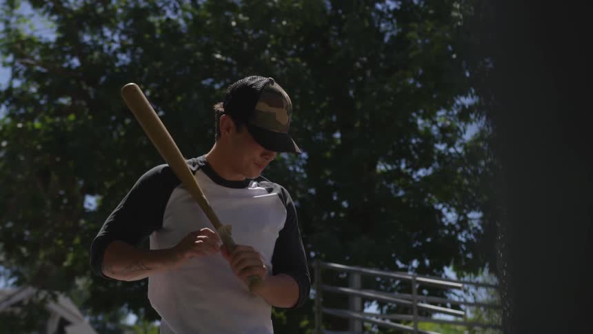 A man stands looking ahead with a baseball bat on his shoulder until he takes a batting stance