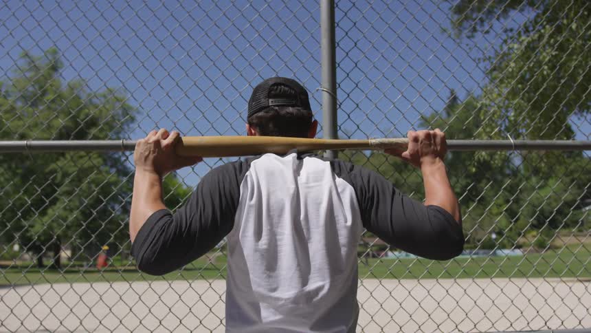 A man looks through the chain link at the infield with a bat resting behind his head on his shoulders